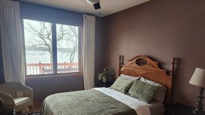 A bedroom with a large bed, a chair, a nightstand, and a window with a view of a red fence and trees.