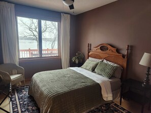 A bedroom with a large bed, a chair, a nightstand, and a window with a view of a red fence and trees. - Luxury Family 5BR Lakefront private boat launch (Big Stone City)