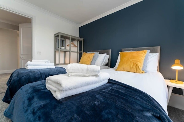 Ein Hotelzimmer mit zwei Betten, jeweils mit einer dunkelblauen Bettdecke und weißen Handtüchern. Ein kleiner Nachttisch mit einer Lampe steht daneben.