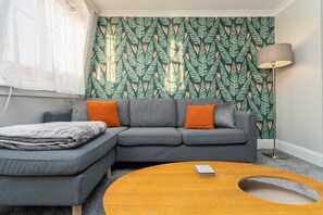 A modern living room with a grey sectional sofa, orange cushions, a wooden coffee table, and a patterned wallpaper.