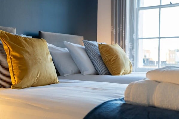 A neatly made bed with multiple pillows and a blanket, a window with curtains, and a view of buildings outside.
