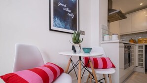 Una cocina moderna con armarios blancos, una estufa y un microondas. Hay una pequeña mesa redonda con un cuenco azul y una planta en maceta. Dos sillas blancas con cojines a rayas rojas y rosadas están situadas frente a la mesa.