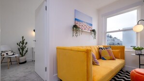Una sala de estar moderna con un sofá amarillo, un cuadro de una playa, una ventana con vistas a edificios y una planta en maceta.
