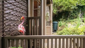 A flamingo standing on a wooden deck with a metal fence and a door leading to a green area with plants.