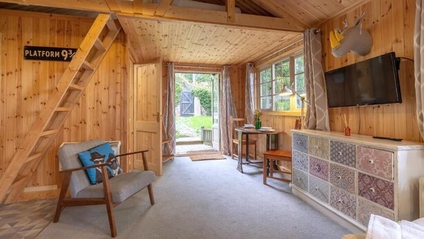 A wooden cabin interior with a staircase, a TV mounted on the wall, a small table with chairs, and a sofa.