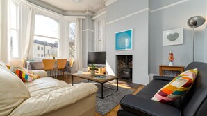 Een moderne woonkamer met een bank, een zwarte fauteuil, een salontafel en een open haard. Er is een televisie op een dressoir, een eettafel met stoelen, en een raam met gordijnen.