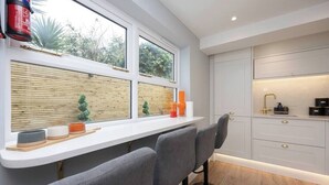 A modern kitchen with a white countertop, two bar stools, and a window with a view of greenery. - Host & Stay | Brighton Laines House (Brighton and Hove)