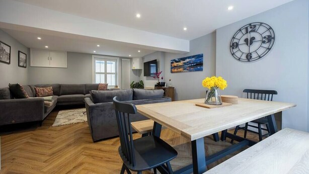 A modern living room with a sofa set, a dining table with chairs, a clock on the wall, and a television mounted above the sofa. - Host & Stay | Brighton Laines House (Brighton and Hove)