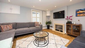 A living room with a grey sofa, a glass coffee table, a fireplace, and a TV mounted on the wall. - Host & Stay | Brighton Laines House (Brighton and Hove)