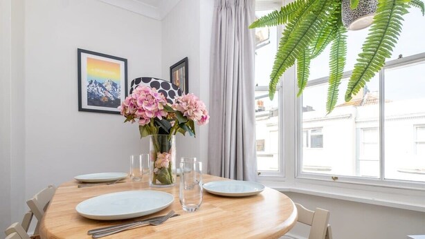 A dining area with a round wooden table, white plates, cutlery, and a vase with pink flowers. There are framed pictures on the wall and a large window with a view of buildings. - Host & Stay | Guildford Apartment (Brighton and Hove)