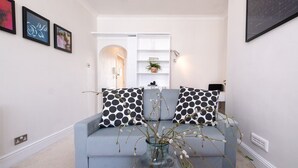 A modern living room with a light blue sofa, black and white polka dot cushions, a glass vase with branches, and a white shelving unit against a white wall. - Host & Stay | Guildford Apartment (Brighton and Hove)