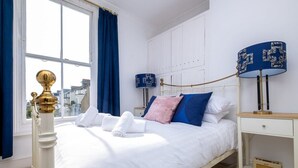 A neatly made bed with white linens and rolled towels, a bedside table with a lamp, and a window with blue curtains. - Host & Stay | Guildford Apartment (Brighton and Hove)