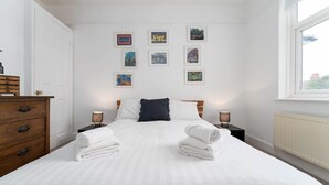 A neatly made bed with white linens and two rolled towels, a wooden dresser, and a wall with framed pictures. - Host & Stay | Preston Park Apartment (Brighton and Hove)