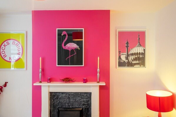 A room with a pink wall, a fireplace, and two framed pictures.