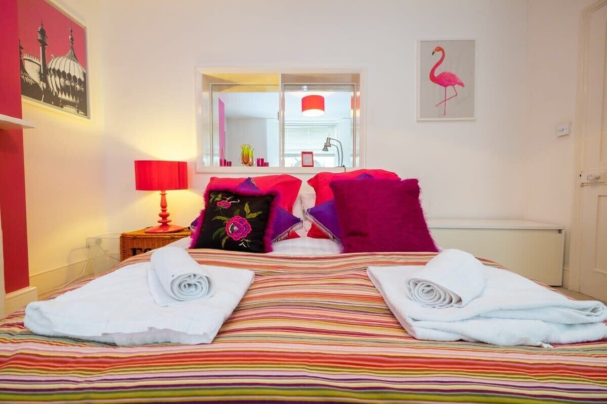A hotel room with a striped bedspread, two rolled towels on the bed, a red lamp on a bedside table, a window with a view of the outside, and a pink flamingo painting on the wall.