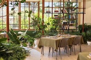 A dining area with a table set for two, surrounded by potted plants, and a bookshelf filled with books. - Le Macaron Garden Stay - Boutique Hotel Da Lat (Cam Ly - Da Lat)