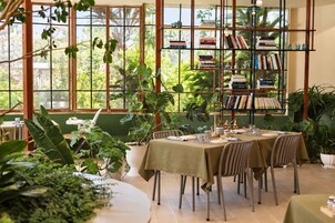 A dining area with a table set for two, surrounded by potted plants, and a bookshelf filled with books.