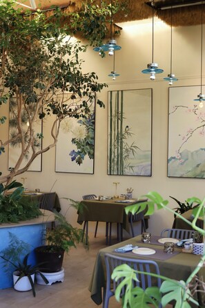 Un espace repas avec des tables préparées pour deux personnes, entouré de plantes en pot et d’œuvres d’art accrochées aux murs.