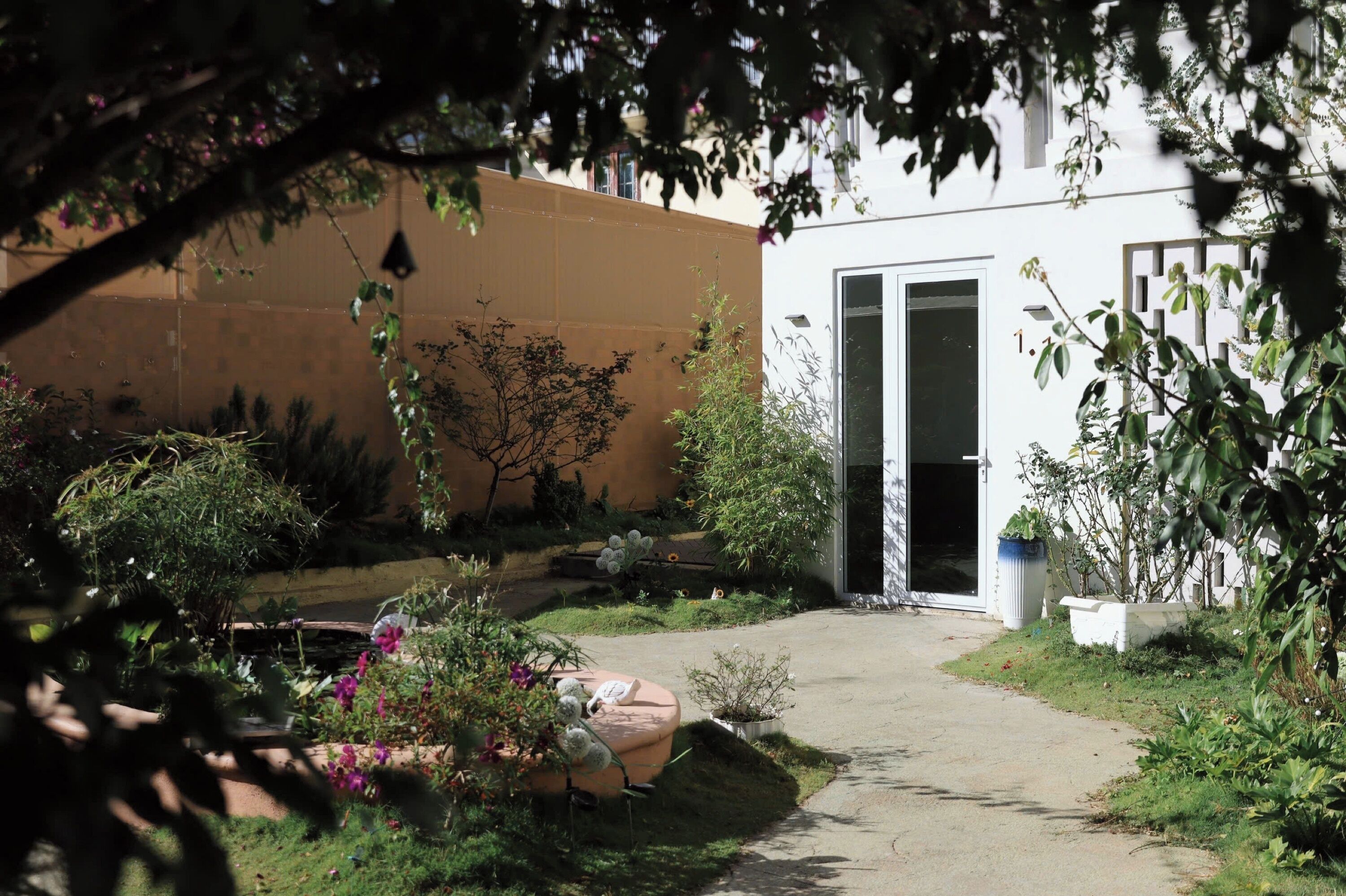 A garden with a pathway leading to a building with a door. There are various plants and a small white structure in the garden.