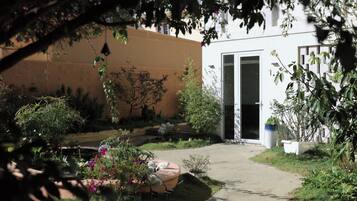 A garden with a pathway leading to a building with a door. There are various plants and a small white structure in the garden.