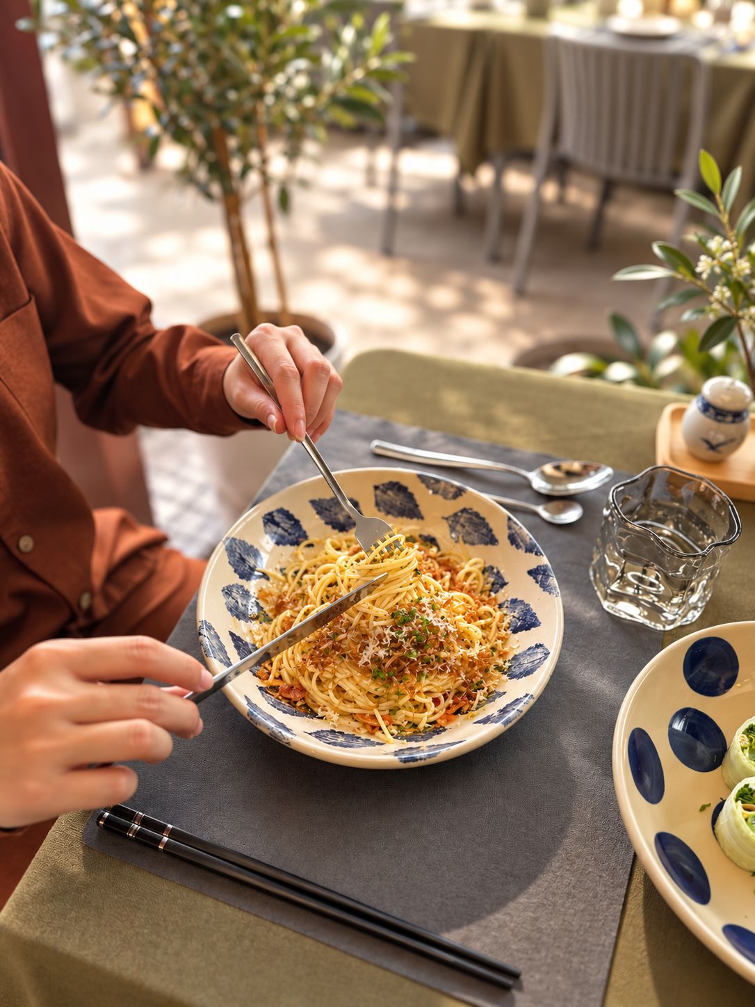 A person dining outdoors with a plate of spaghetti, a glass of water, and chopsticks on a table.