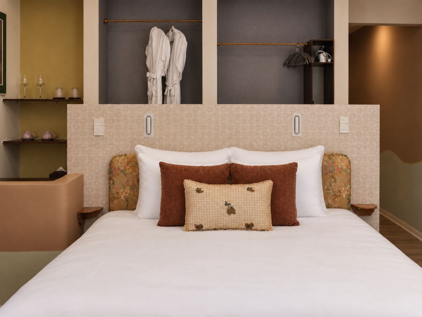 A hotel room with a bed, two bedside tables, a wardrobe, and a shelf with toiletries.
