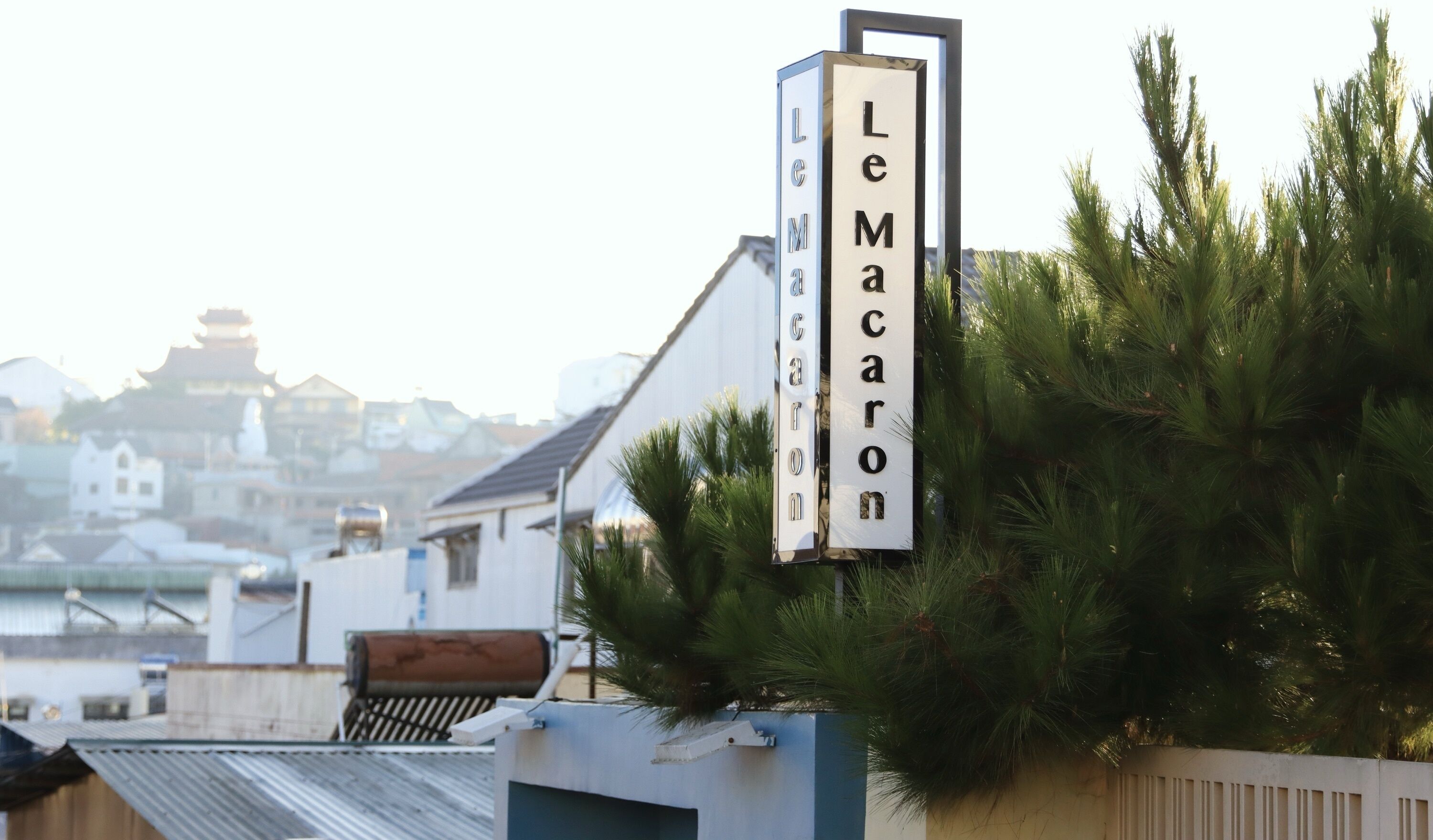 A sign with the name 'Le Macaron' in a vertical format, mounted on a structure with a clear view of a traditional building and a pagoda in the background.