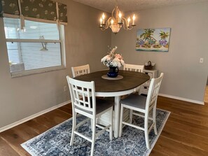 A dining room with a round table, white chairs, a chandelier, a painting of a tropical scene, and a window with blinds.