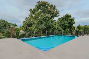 Ein rechteckiger Swimmingpool mit blauer Umrandung, umgeben von einem Metallzaun und Bäumen im Hintergrund.