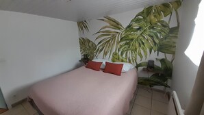 Un dormitorio con una cama, dos almohadas rojas, un protector de colchón blanco y una planta grande en maceta contra la pared.