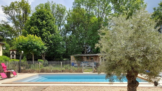 Una piscina con cubierta de madera, rodeada de piedras y rocas. Se ven sillas de descanso rosadas y una valla de madera al fondo.