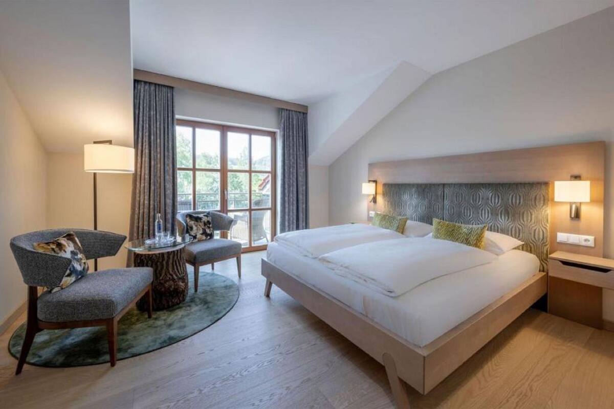 Una habitación de hotel moderna con una cama grande, dos mesitas de noche, una silla, una pequeña mesa redonda y un ventanal con cortinas.