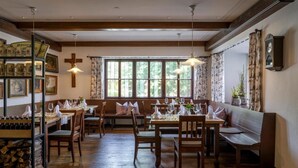 Un coin repas chaleureux avec des tables et des bancs en bois, une croix au mur et une grande fenêtre avec des rideaux.