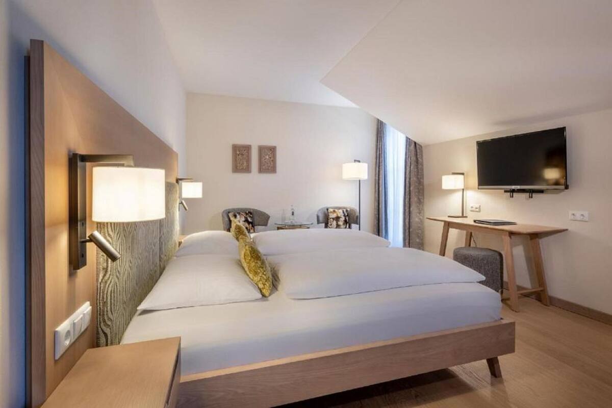 Habitación de hotel moderna con una cama grande, lámparas de noche, un escritorio con una silla, un televisor de pantalla plana y una ventana con cortinas.
