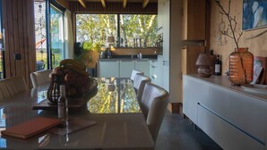 Una cocina moderna con un amplio ventanal, armarios de madera y una zona de comedor con mesa y sillas de vidrio.