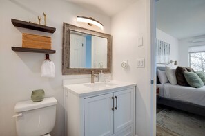 Ein Badezimmer mit einem weißen Waschbecken, einem Spiegel und einer Toilette. Über dem Waschbecken befinden sich Regale mit dekorativen Gegenständen.