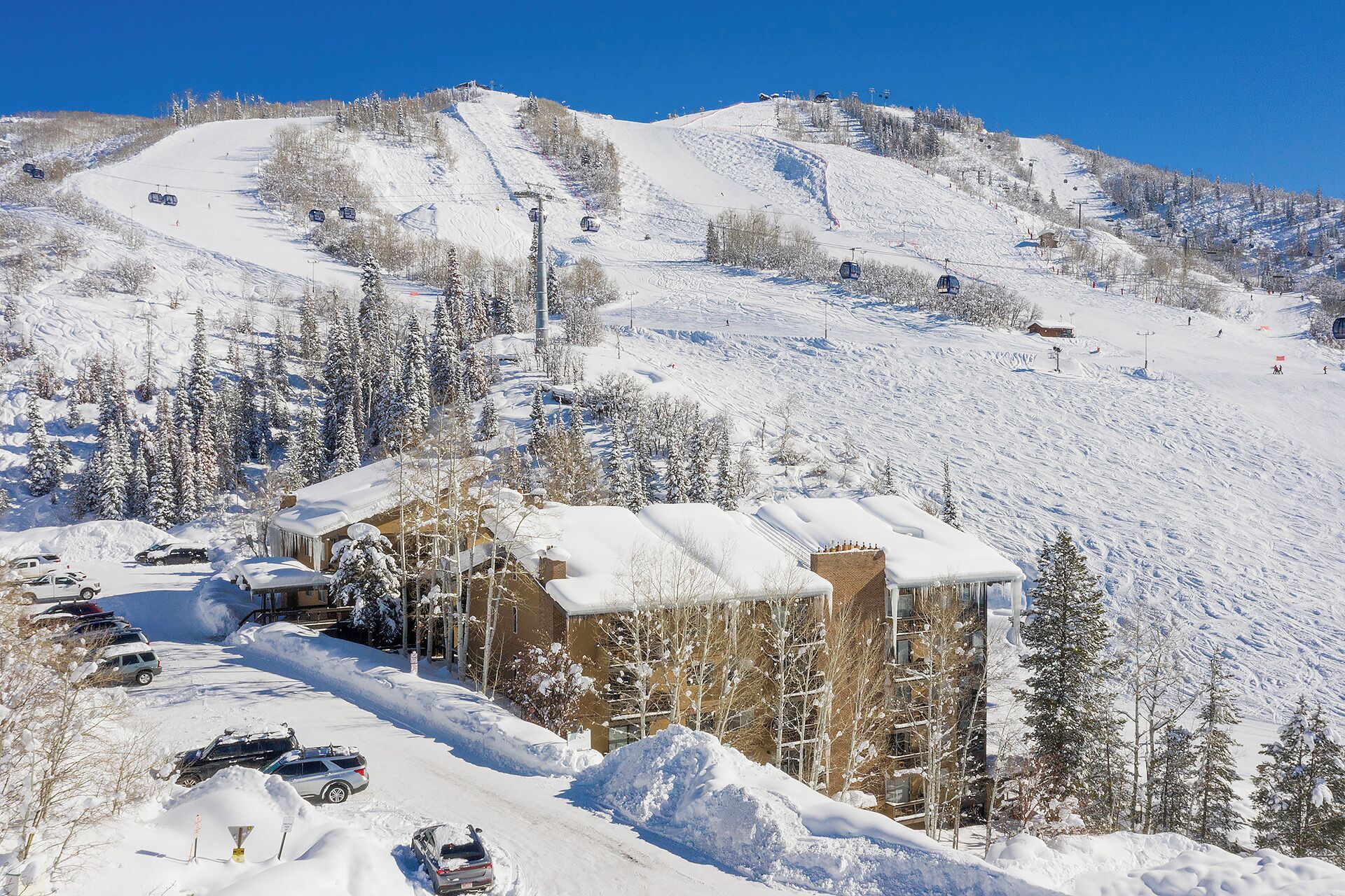 A snow-covered mountain with a ski lift, a building with a snow-covered roof, and parked cars.