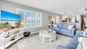 A modern living room with a large flat-screen TV, a blue sofa, a white coffee table, and a wicker side table.