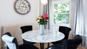 Un'area pranzo con un tavolo rotondo in marmo, sedie rivestite in tessuto nero, un vaso con fiori, una sveglia a muro e una finestra con tende.