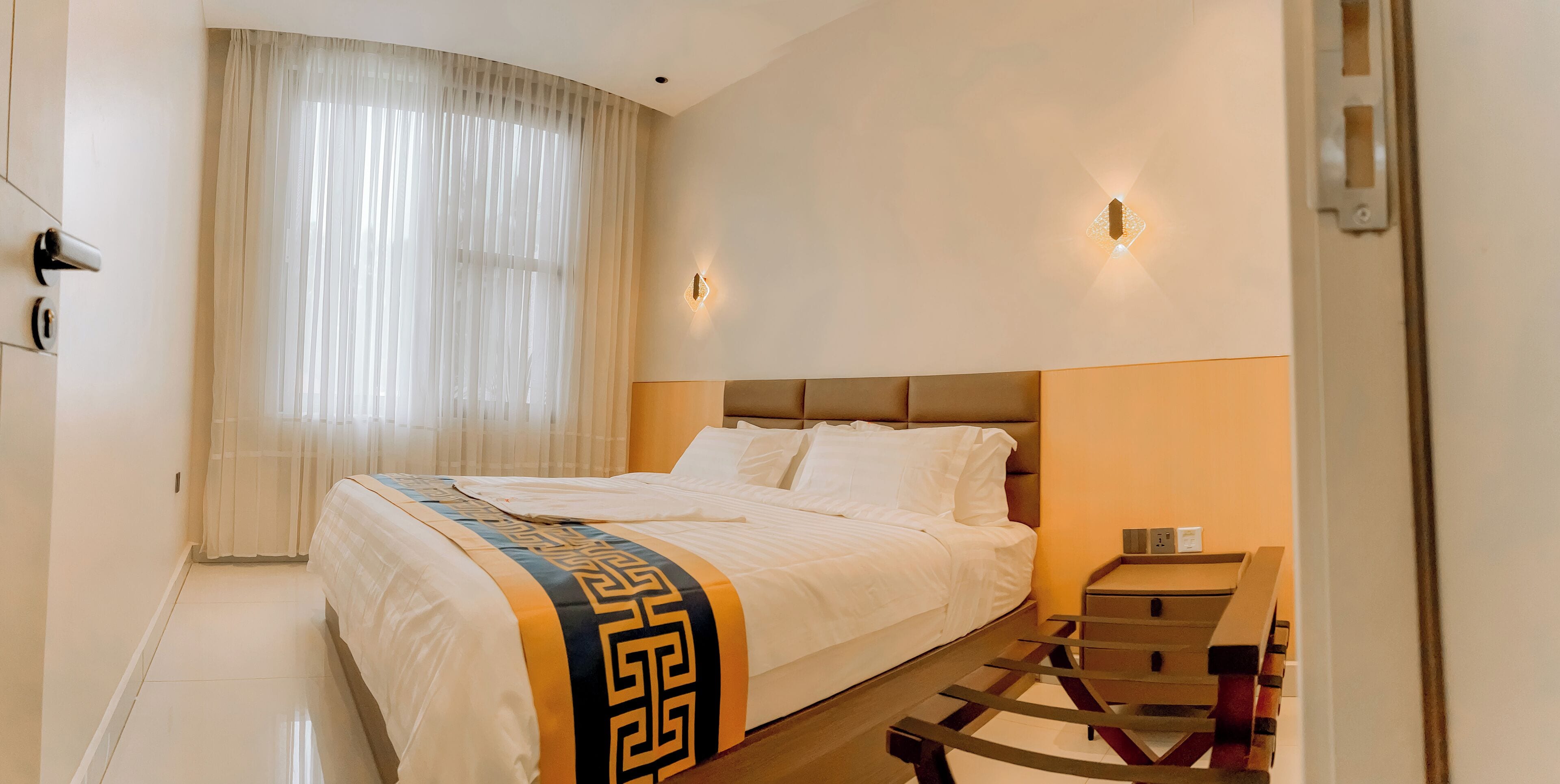 A hotel room with a bed, a nightstand, a lamp, and a window with sheer curtains.