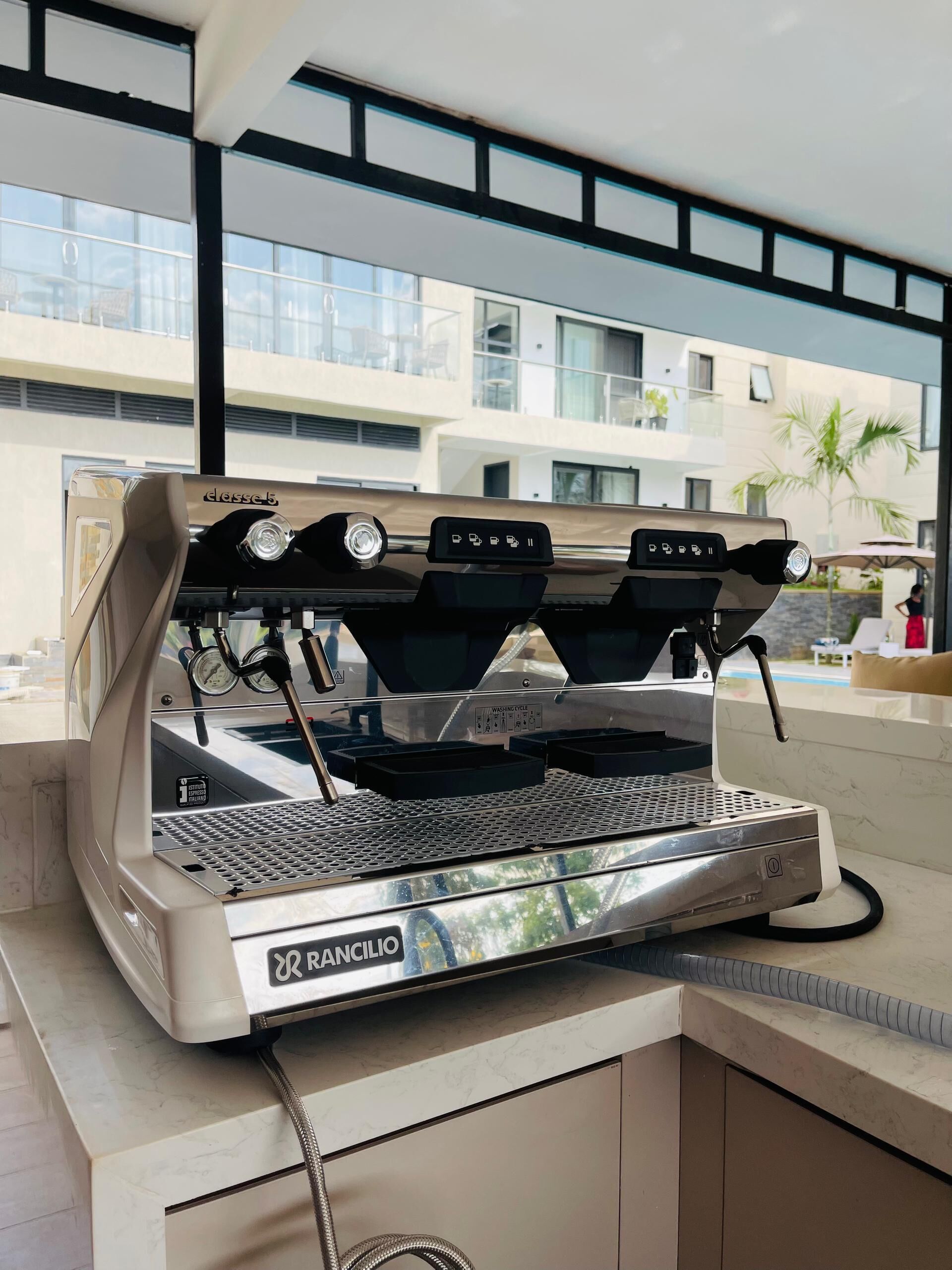 A Rancilio espresso machine in a modern kitchen with a view of a building and palm trees outside.