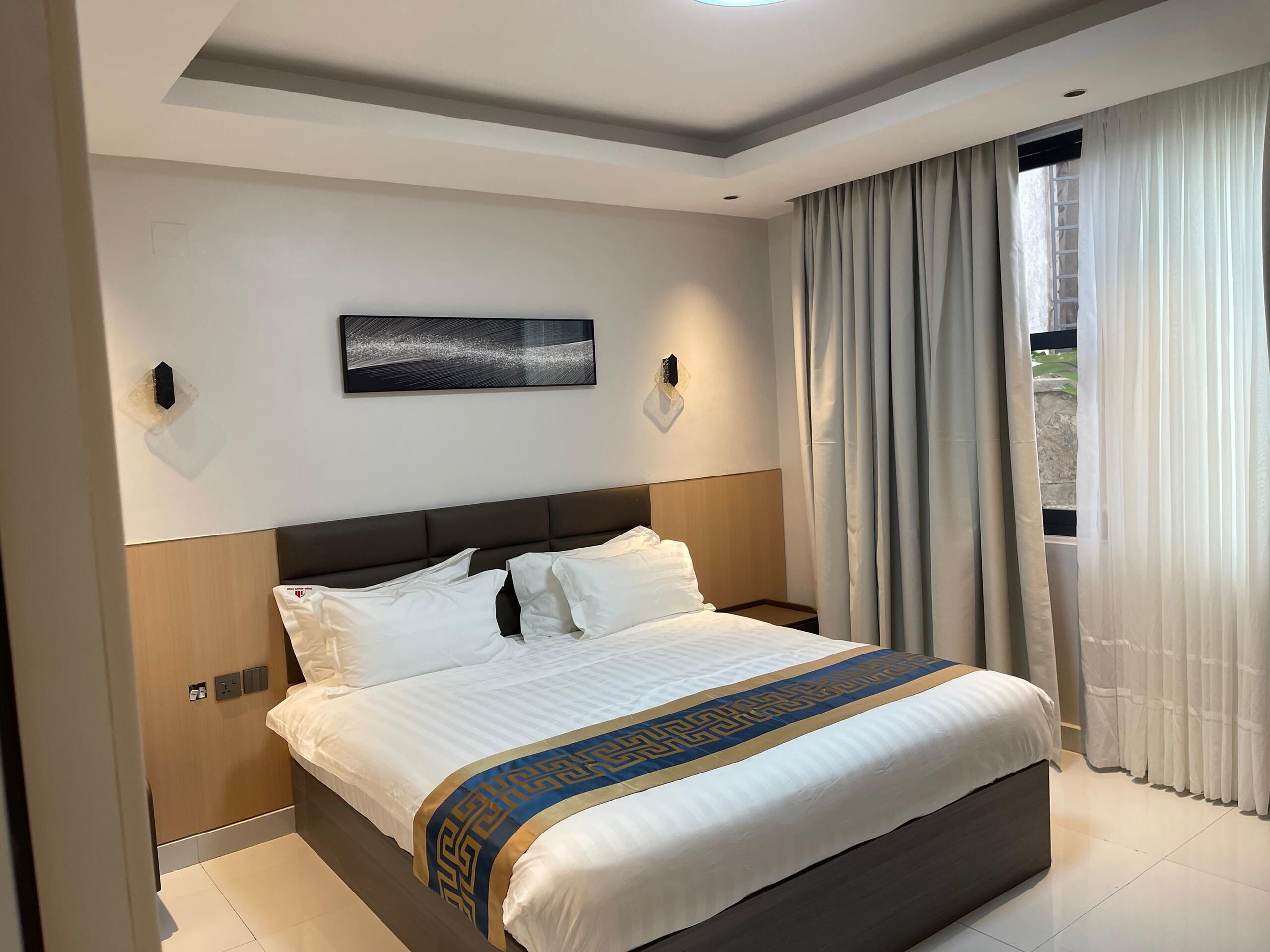 Habitación de hotel con una cama grande, cabecera, una obra de arte colgada en la pared y una ventana con cortinas.