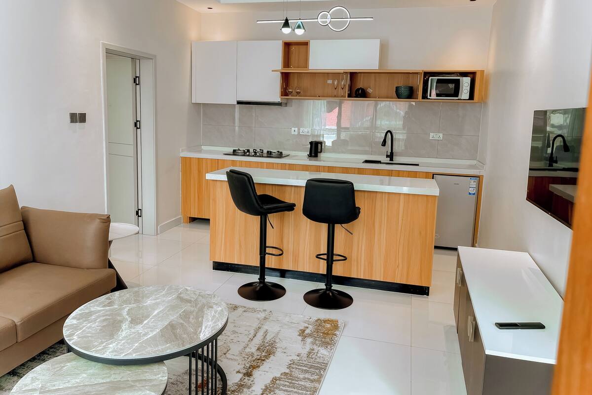A modern kitchen with a central island, two black bar stools, a white countertop, and a built-in microwave.