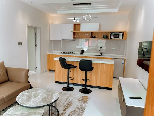 A modern kitchen with a central island, two black bar stools, a white countertop, and a built-in microwave. - Step Apart Hotel (Kigali)