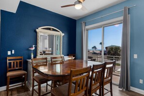 Un espace repas comprenant une grande table et des chaises, un ventilateur de plafond et une vue sur l’extérieur grâce à de grandes fenêtres.