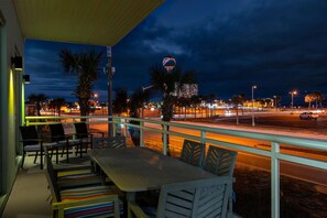 Un balcon avec des chaises et une table, donnant sur une rue bordée de palmiers et sur un bâtiment illuminé en arrière-plan.