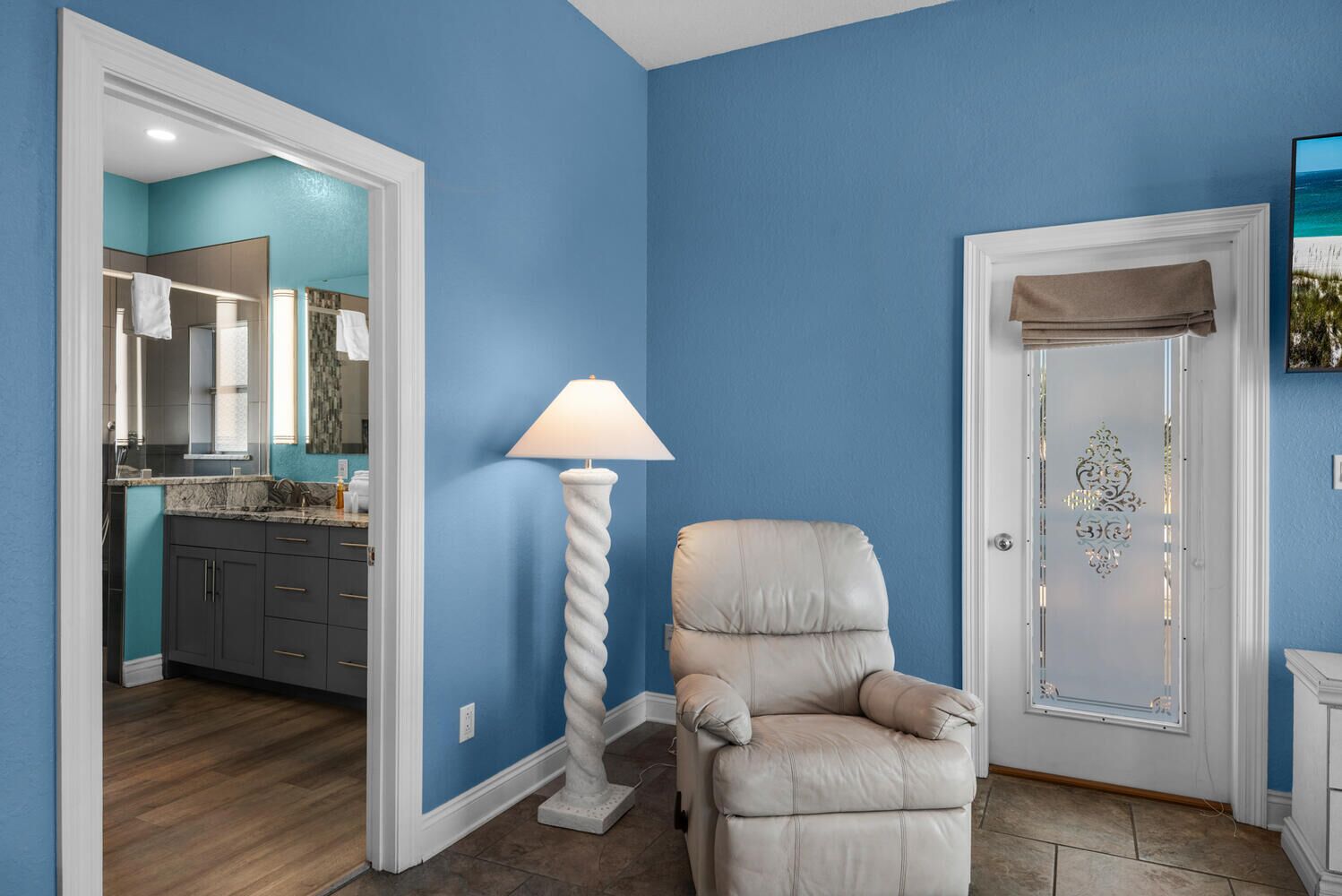 Una habitación con pared azul, un sillón color beige, una lámpara de pie blanca y una puerta con un panel de vidrio decorativo.