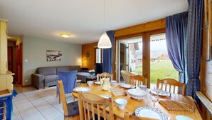 Een eetgedeelte met een houten tafel voor vier personen, een bank en uitzicht op een woonwijk door een glazen deur.