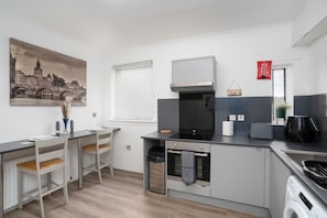 A modern kitchen with grey cabinets, a built-in oven, a microwave, and a washing machine. There is a window with blinds, a painting of a cityscape, and a red fire safety sign. - 2-bed Flat, Wishaw, Free Parking, Family-friendly (Wishaw)