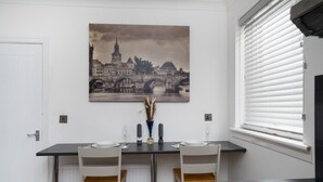 A dining area with a table set for two, a cityscape painting above, and a window with blinds. - 2-bed Flat, Wishaw, Free Parking, Family-friendly (Wishaw)